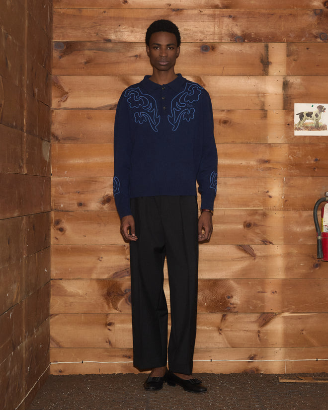 A person wearing the Blaze Polo - Navy, which features subtle Western floral patterns, paired with black pants, stands against a wooden wall; a small dog photo and a fire extinguisher are also visible.