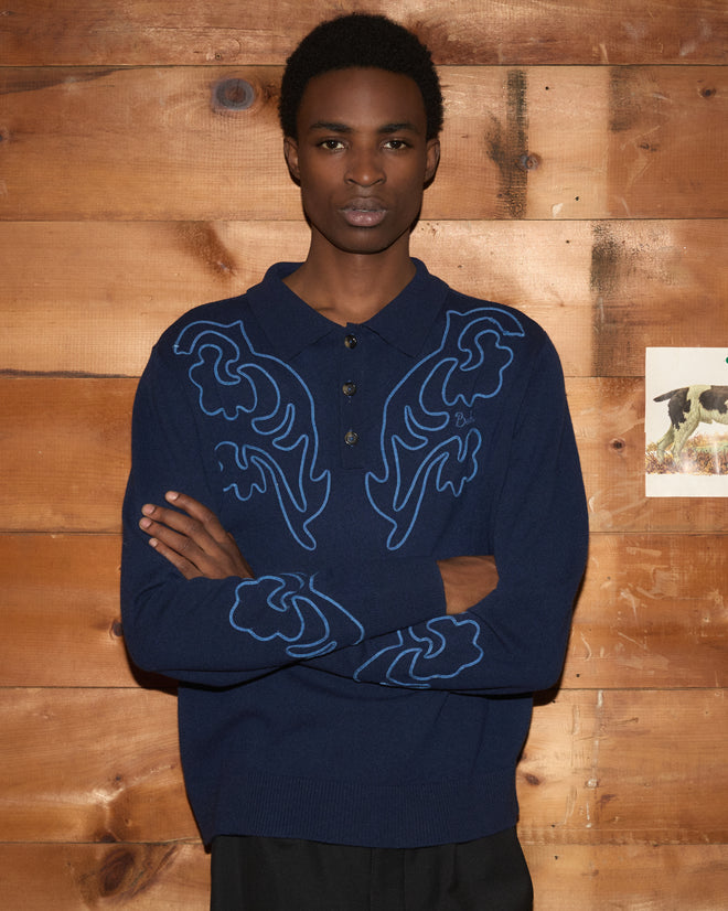 Wearing the Blaze Polo - Navy, which features blue Western floral patterns, a person stands with arms crossed in front of a wooden wall.