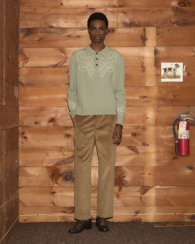 Wearing the Blaze Polo in Sage, a person in tan pants stands in a wooden room near a fire extinguisher.