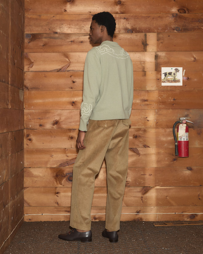 A person stands in a wood-paneled room, facing away, wearing the Blaze Polo in Sage near a fire extinguisher and a small picture on the wall.