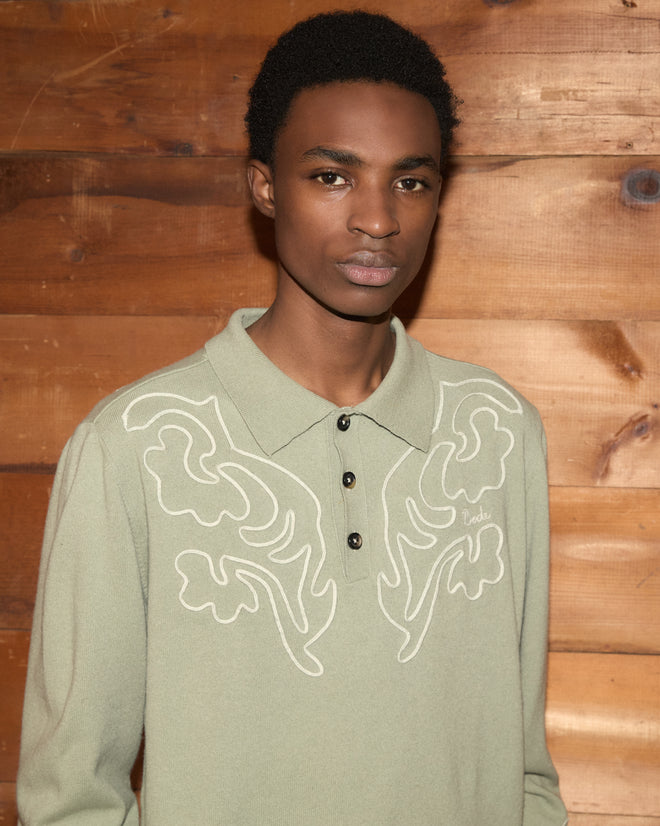 A person wearing the Blaze Polo - Sage, featuring white embroidery and subtle Western floral patterns, stands in front of a wooden wall, looking at the camera.