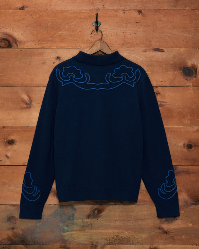 Blaze Polo - Navy featuring blue Western floral patterns and delicate embroidery on the back and sleeves, accented with subtle soutache braid details, displayed against a wooden wall.
