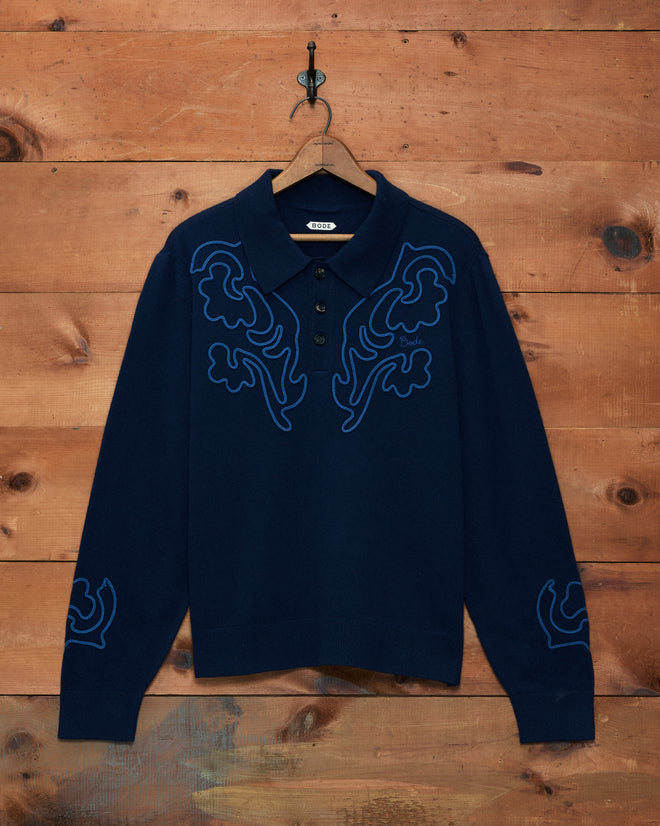 Blaze Polo - Navy: A long-sleeve navy blue sweater featuring blue abstract embroidery with subtle hints of Western floral patterns, displayed against a wooden wall.