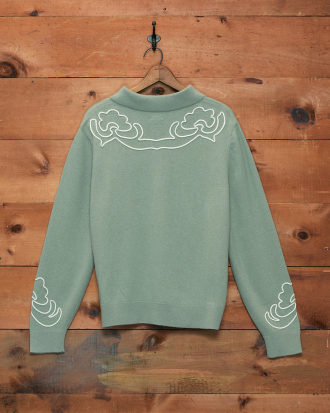 The Blaze Polo - Sage, featuring light sage green fabric adorned with white Western floral patterns, hangs on a hook against a wooden wall.