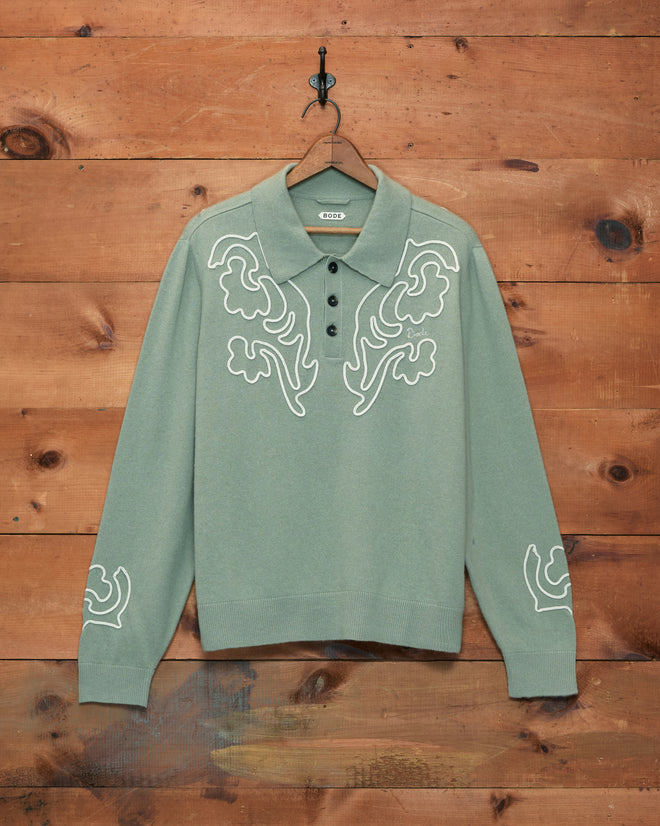 Blaze Polo - Sage, a long-sleeve sage green sweater featuring white embroidered Western floral patterns, displayed hanging on a wooden wall.