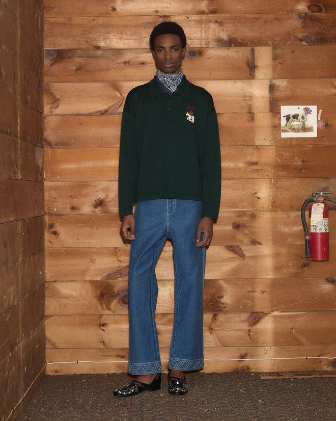 A person stands in front of a wooden wall, wearing jeans, the Cowboy Appliqué Polo - Green, and black shoes.