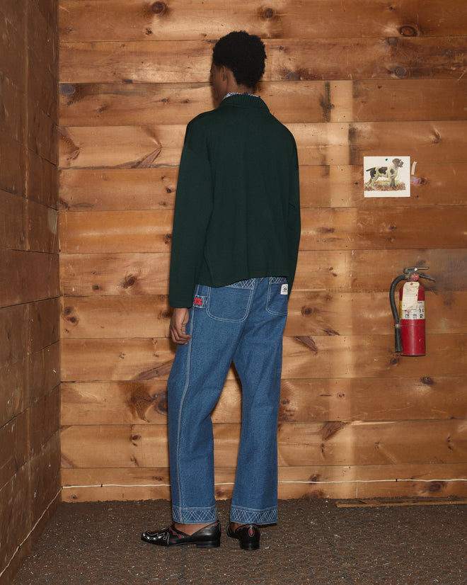 A person stands with their back to a wooden wall, dressed in jeans, black shoes, and a green Cowboy Appliqué Polo featuring a charming, hand-embroidered felt pony on the back.