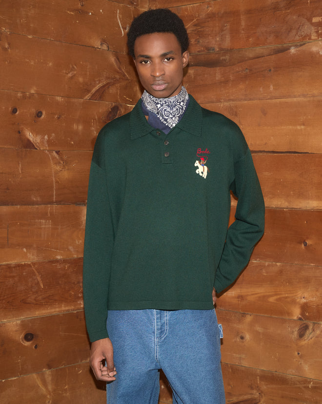 A person wearing the Cowboy Appliqué Polo in green, paired with jeans and a blue bandana, stands against a wooden wall.