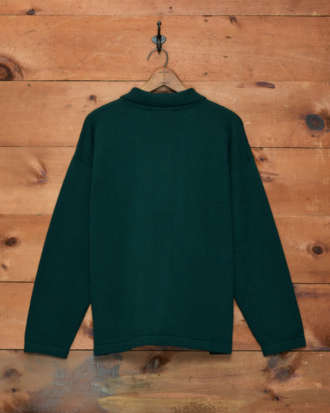 Cowboy Appliqué Polo - Green merino wool sweater hanging on a wooden hanger against a rustic wooden wall background.