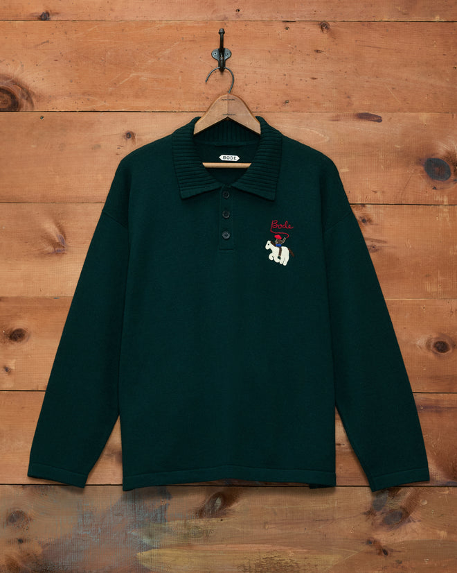 Cowboy Appliqué Polo - Green, a dark green long-sleeve merino wool polo shirt with a bear and “Bode” hand-embroidered on the chest, hanging on a wooden wall.