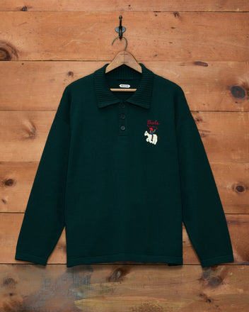 Cowboy Appliqué Polo - Green, a dark green long-sleeve merino wool polo shirt with a bear and “Bode” hand-embroidered on the chest, hanging on a wooden wall.