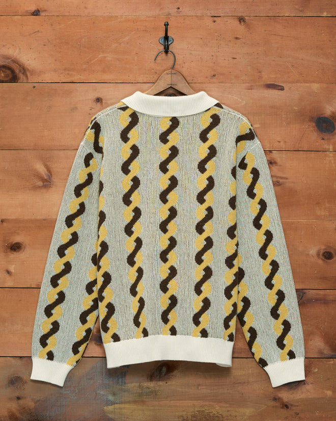 Lasso Twist Polo featuring yellow, brown, and gray wavy stripes in a patterned knit, crafted from soft merino wool and baby alpaca, displayed against a wooden wall.