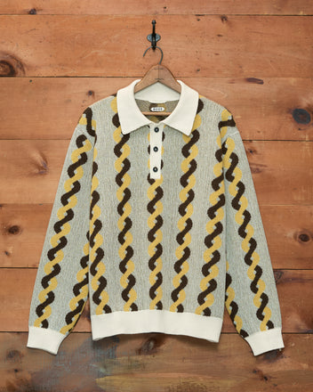 The Lasso Twist Polo, a long-sleeve knit jacquard pullover featuring a collared neck and crafted from soft merino wool, is displayed hanging on a wooden wall.
