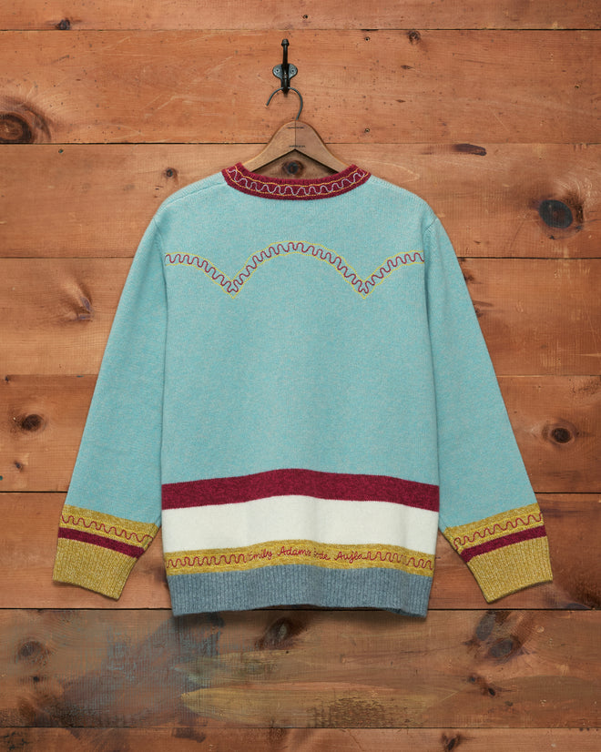 The Rock Candy Cardigan, featuring a classic Western yoke detail, hangs on a wooden wall, its blue fabric accented by bold red and yellow trim.