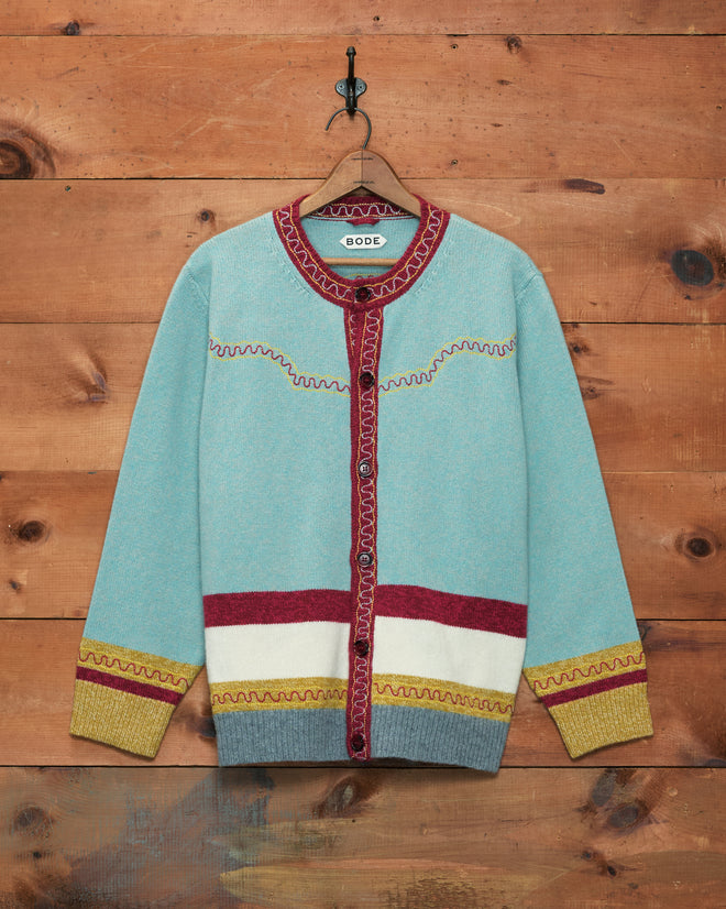 A light blue Rock Candy Cardigan with red trim, yellow cuffs, and a white band, featuring a classic Western yoke detail and crafted from 100% wool, hanging on a wooden wall.