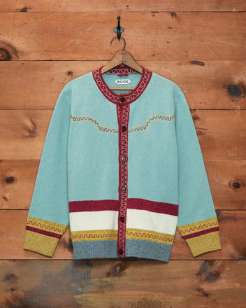 A light blue Rock Candy Cardigan with red trim, yellow cuffs, and a white band, featuring a classic Western yoke detail and crafted from 100% wool, hanging on a wooden wall.