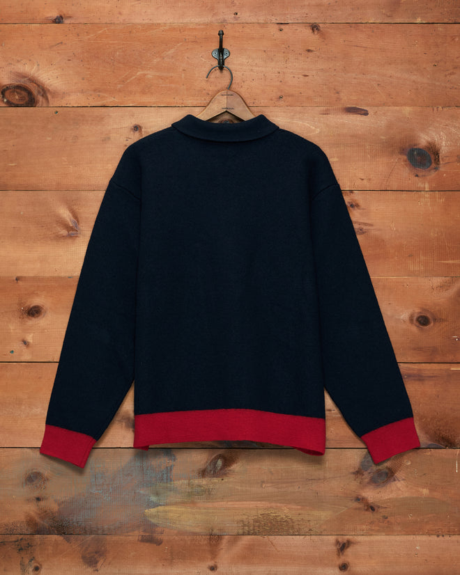The Mr. Checkers Felted Pullover, featuring a navy blue design with red cuffs and hem, is displayed on a wooden wall on a hanger and shown from the back.