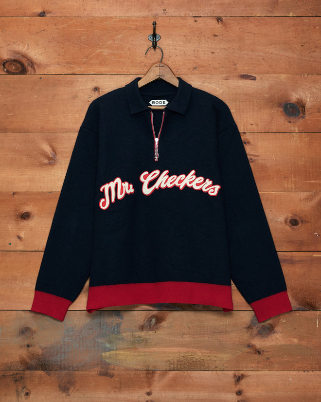 The Mr. Checkers Felted Pullover—crafted from 100% wool—features a black design with red cuffs and hem, “Mr. Checkers” text on the front, and is shown hanging on a wooden wall for a stylish rodeo-inspired look.