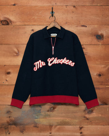 The Mr. Checkers Felted Pullover—crafted from 100% wool—features a black design with red cuffs and hem, “Mr. Checkers” text on the front, and is shown hanging on a wooden wall for a stylish rodeo-inspired look.