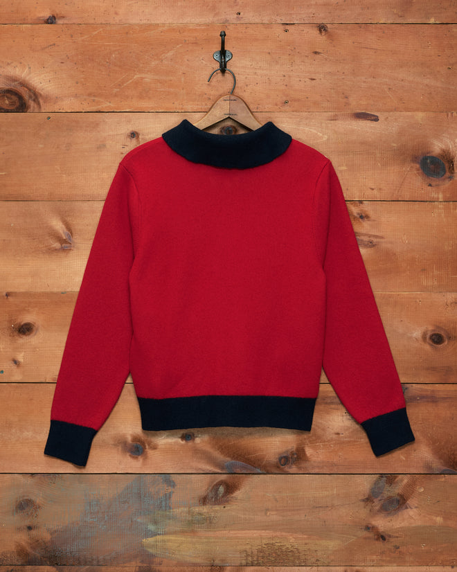 The Mr. Checkers Felted Cardigan, a red long-sleeve zip sweater with black collar and cuffs, crafted from 100% wool, hangs on a hook against a wooden wall.