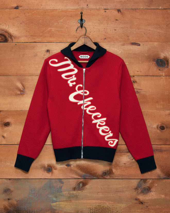 The Mr. Checkers Felted Cardigan features a red design with black collar and cuffs, expertly crafted from 100% wool. “Mr. Checkers” is displayed diagonally across the front, combining classic rodeo knitwear inspiration with a contemporary zip-up cardigan silhouette.