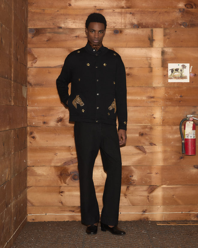 A person in black clothing, possibly wearing the Star Pony Cardigan from the latest men’s knitwear collection, stands in front of a wooden wall adorned with a fire extinguisher and a small animal photo.