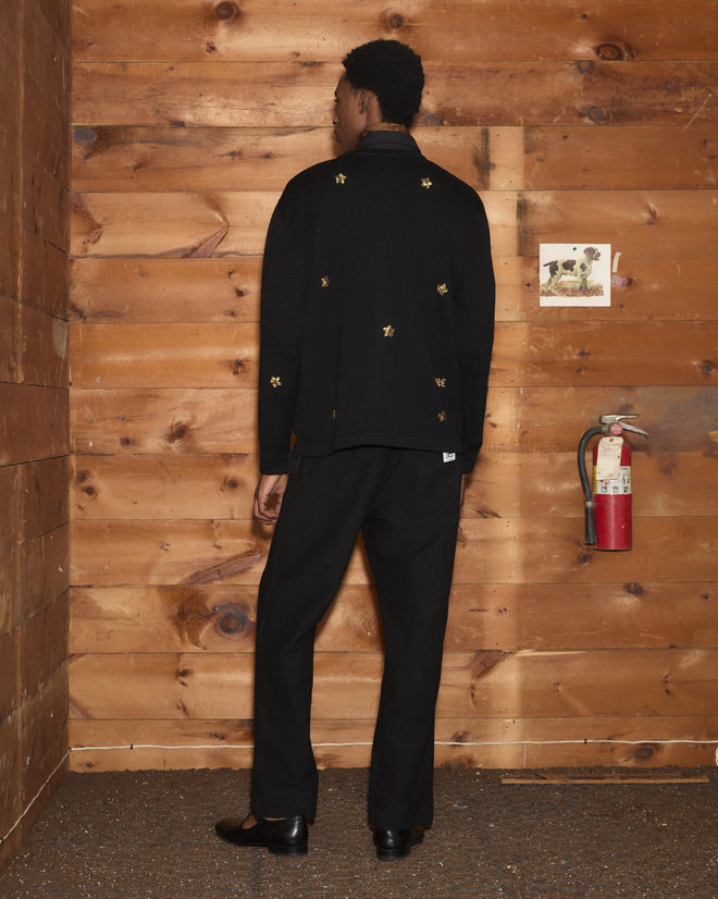 Wearing the Star Pony Cardigan in black, the person stands facing a wooden wall, with a fire extinguisher and a dog photo visible nearby.