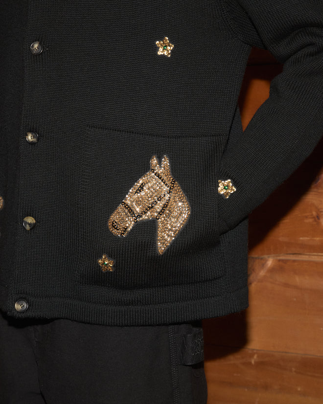 The Star Pony Cardigan, crafted in black virgin wool and adorned with gold horse and flower embellishments, is presented against a wooden background as a refined addition to men’s knitwear collections.