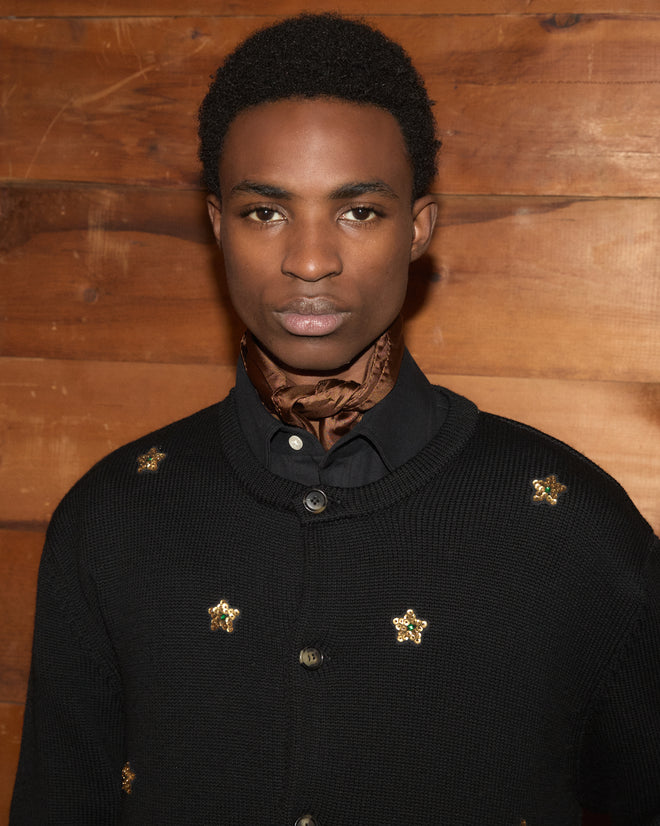A person wearing the Star Pony Cardigan, a black virgin wool piece with gold accents, stands in front of a wooden wall and looks at the camera.