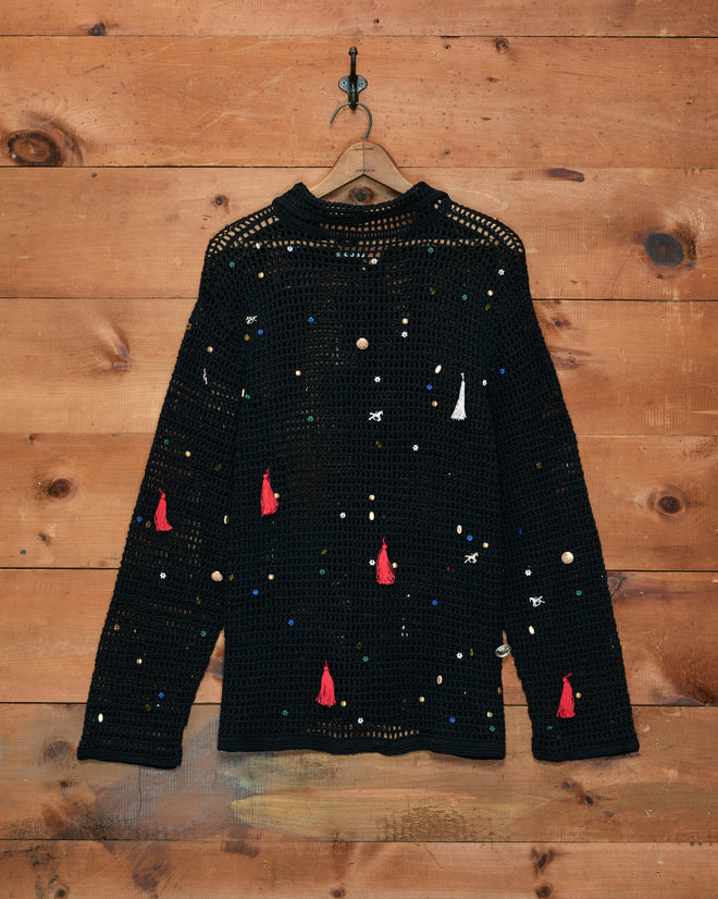 The Crochet Charm Shirt is a black, long sleeve top crafted from 100% cotton, adorned with hand-applied colorful beads and red tassels, and displayed on a wooden wall.