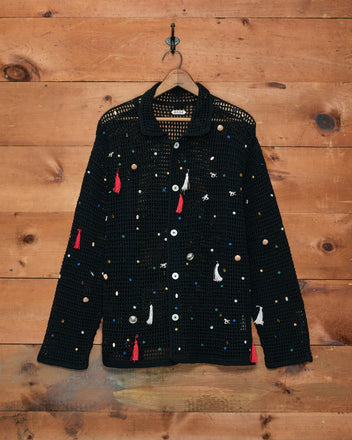 Crochet Charm Shirt—black crocheted shirt adorned with colorful tassels, buttons, and hand-applied embellishments, displayed hanging from a hook on a wooden wall.
