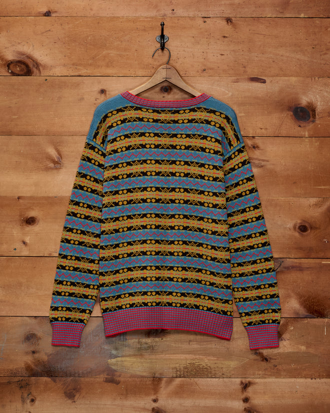 The Bracken Sweater - Blue/Multi, a colorful Fair Isle style knit in blue, yellow, orange, and red and crafted from 100% cotton, is displayed on a hanger against a wooden wall.