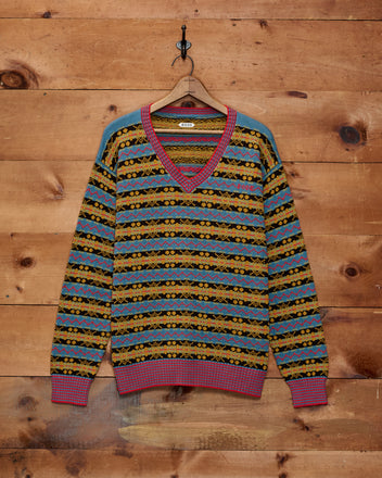 The Bracken Sweater - Blue/Multi, featuring a classic Fair Isle pattern and V-neck design, hangs on a wooden wall by a hanger, highlighting Bode’s signature knitwear craftsmanship.