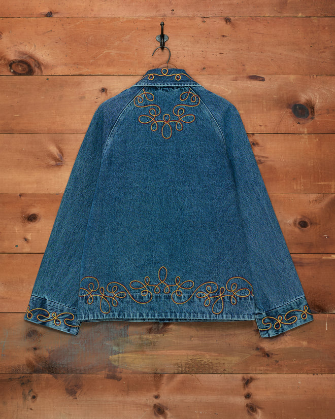 The Rawlins Jacket, a blue denim cowboy-style piece with yellow decorative embroidery, hangs on a wooden wall from a black hook.