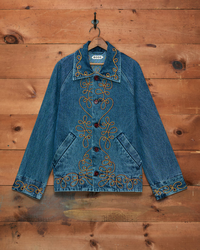 The Rawlins Jacket is a blue denim piece featuring embroidered yellow patterns, displayed on a wooden wall with a black metal hook—an ideal children’s Western jacket for stylish cowboy outfits.