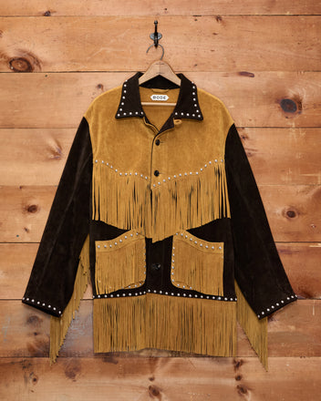 Studded Appalachian Jacket - Brown/Tan: This brown and tan suede fringe cowboy jacket with silver studs hangs on a wooden wall—perfect for a children’s cowboy costume or anyone seeking authentic Western flair.