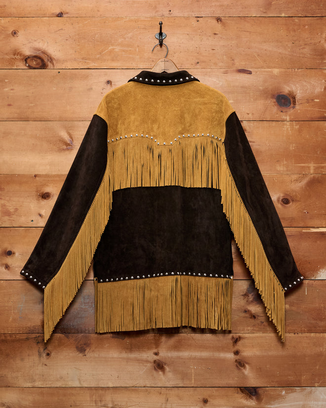 The Studded Appalachian Jacket - Brown/Tan, featuring brown suede with tan fringe and silver studs, hangs on a wooden wall with a metal hook—a perfect addition to any children’s cowboy costume.