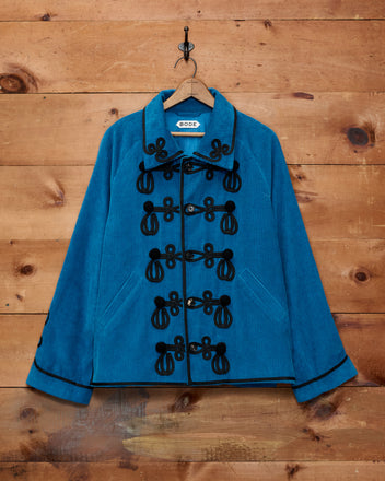 The Stagecoach Corduroy Jacket, featuring a bright blue hue with black soutache braid and crocheted buttons, hangs on a wooden background.
