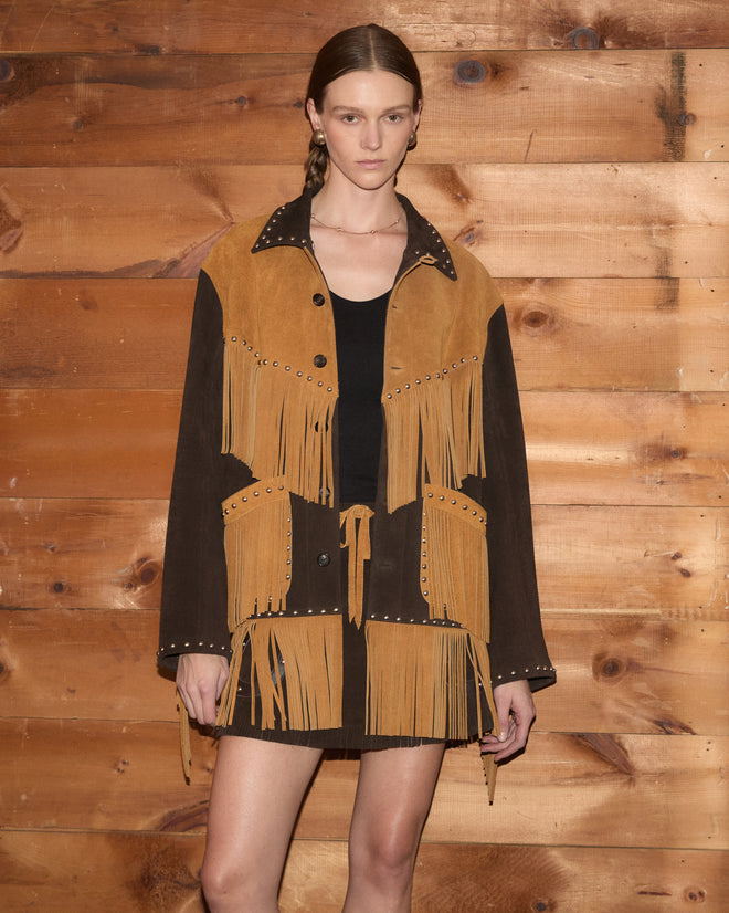A woman in a black dress wears the Studded Appalachian Jacket in brown/tan as she stands against a wooden wall, looking at the camera.