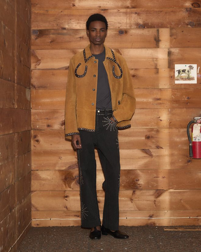 A person in a Suede Carson Jacket and black pants stands in front of a wooden wall with a fire extinguisher on the right.