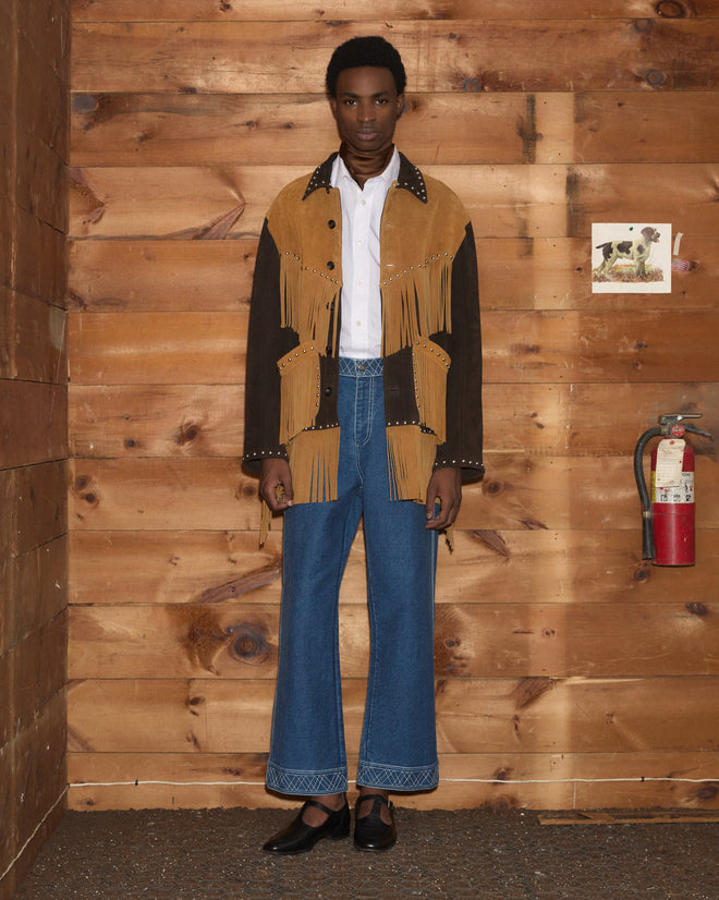 A person wearing the Studded Appalachian Jacket - Brown/Tan and flared jeans stands indoors by a wood wall, fire extinguisher, and art.