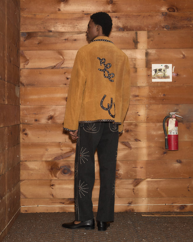 A person stands indoors, facing away, wearing the Suede Carson Jacket in tan and black pants with white patterns, positioned near a fire extinguisher.