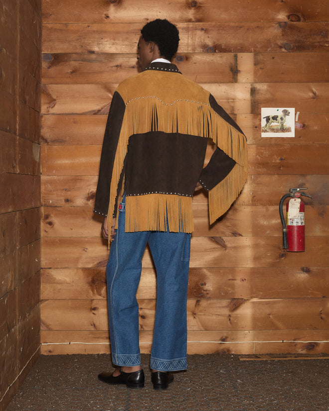 Wearing the Studded Appalachian Jacket in Brown/Tan and blue jeans, a person stands indoors near a wood wall and a fire extinguisher.