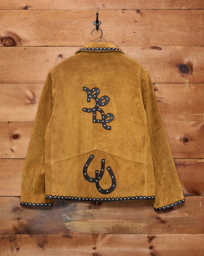 The Suede Carson Jacket in tan, featuring “Be Do” and horseshoe designs on the back, hangs on a wooden wall.