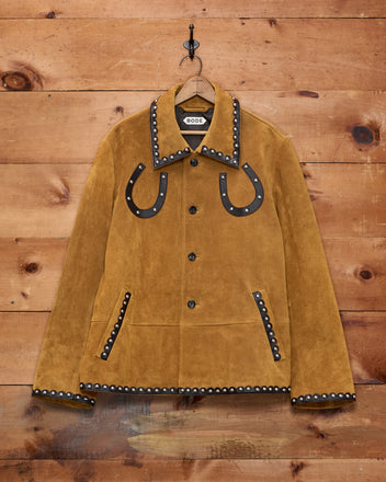 The Suede Carson Jacket is a tan suede outerwear piece with black studded trim and classic horseshoe detailing on the chest, displayed against a wooden wall—an iconic Western jacket that blends rugged charm with timeless style.