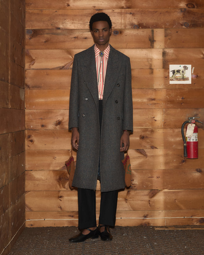 A person wearing a grey Buffalo Tweed coat over a striped shirt with a bolo tie and dark trousers.
