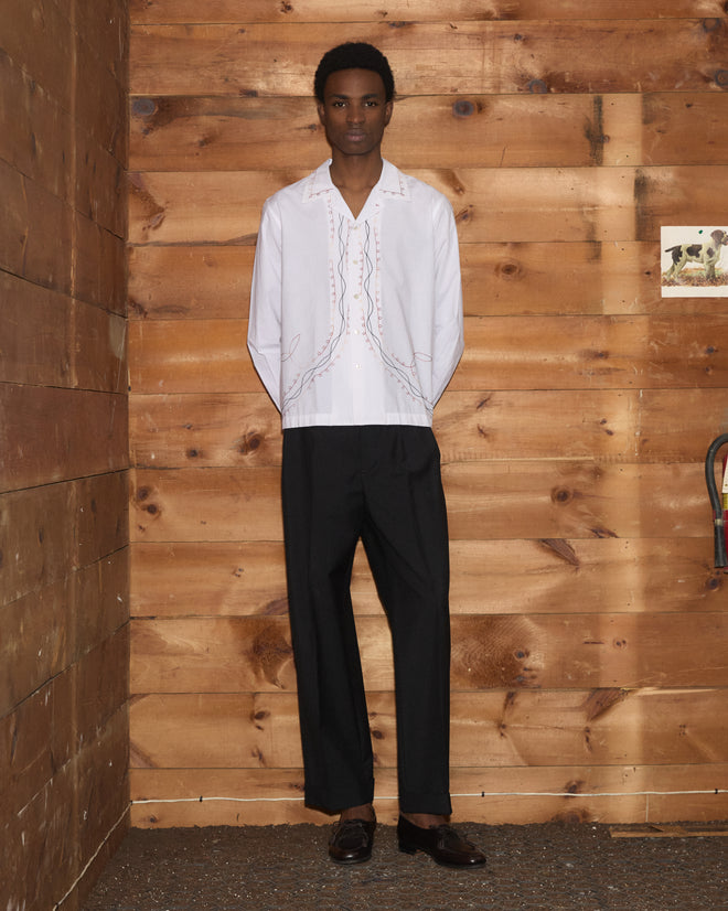 A person stands indoors by a wooden wall, hands behind their back, wearing the Looped Chainstitch Long Sleeve Shirt in white premium cotton poplin and black pants.