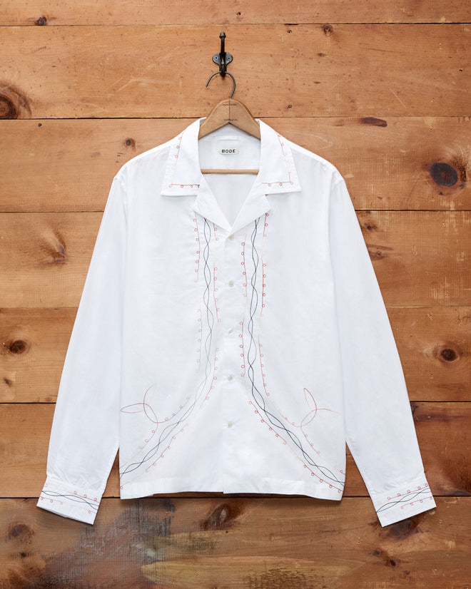 Looped Chainstitch Long Sleeve Shirt, a white cotton poplin button-up featuring delicate pink and blue embroidered lines, crafted in India and displayed on a wooden wall.