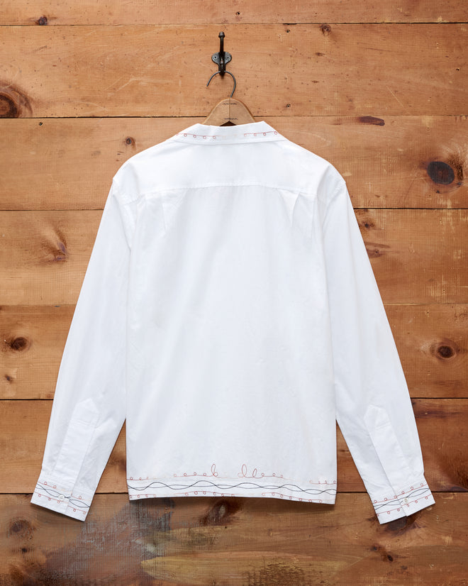 The Looped Chainstitch Long Sleeve Shirt, crafted from soft white cotton poplin with bold red chainstitch detailing, is displayed from the back as it hangs on a wooden wall.
