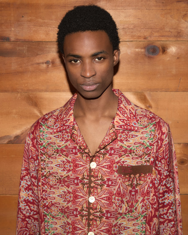 A person wearing the Arrowleaf Long Sleeve Shirt—a red wool top featuring a subtle paisley print—stands in front of a wooden wall, looking directly at the camera.